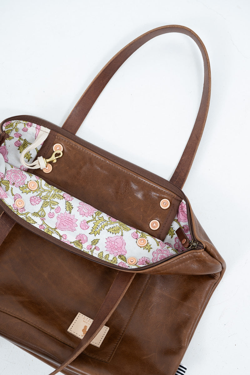A brown leather tote bag shown open from a top-down angle, revealing a floral-patterned interior lining with pink flowers, green leaves, and a white background. The bag has dual shoulder straps, gold-tone hardware, and a small beige brand patch near the bottom.