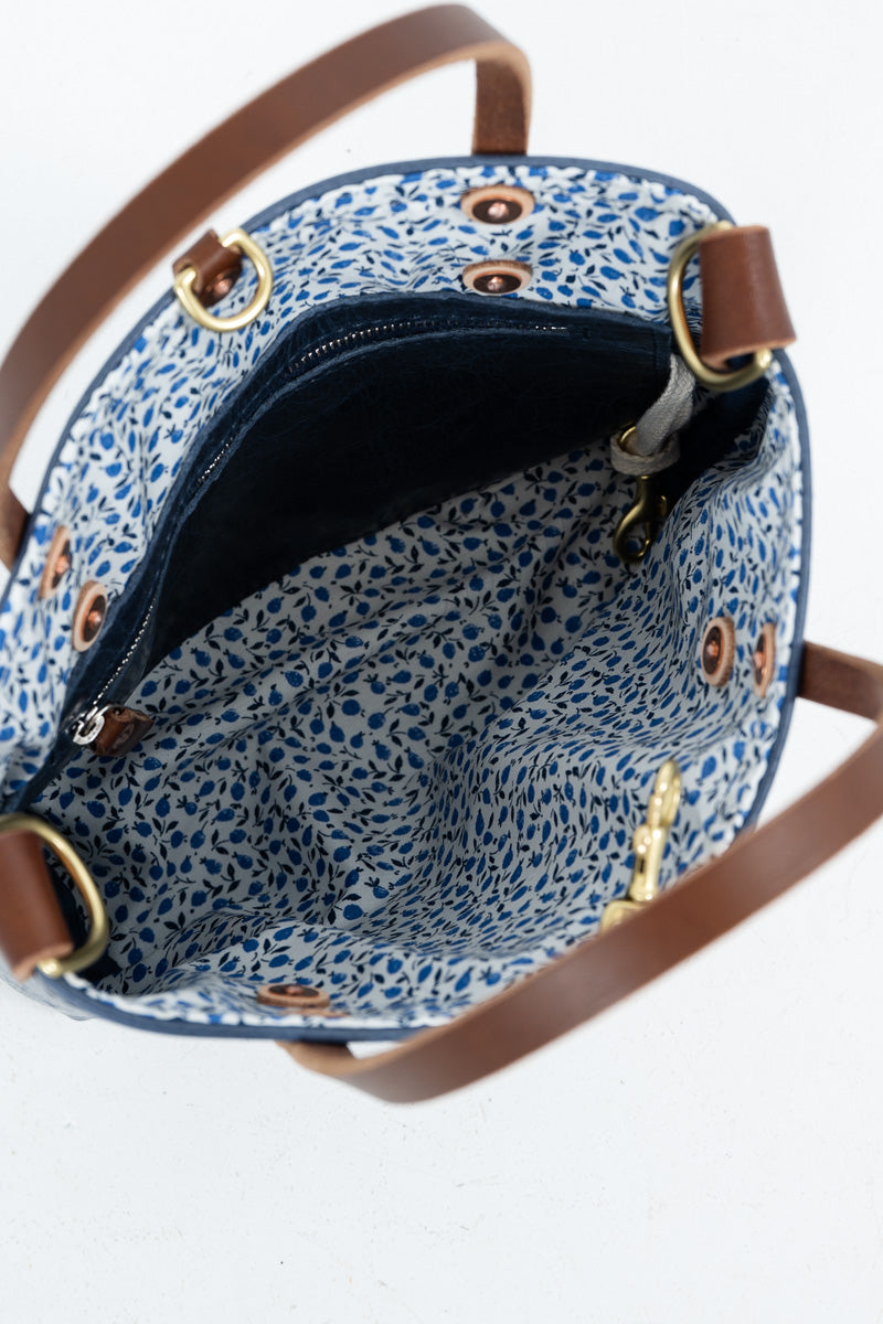 Close-up of the interior of a handbag with a blue and white floral patterned fabric lining, brown leather trim, and brass-tone hardware on a white background.