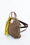 A brown waxed canvas backpack with dark brown leather handles and trim, featuring a saffron yellow polka dot scarf tied to the handle, photographed on a white background. Handmade by Meanwhile Back on the Farm in Lynchburg,VA