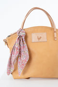 A tan leather handbag with a floral pink scarf tied to the handle, shown against a plain white background. The bag features a rectangular tan leather patch on the front with a rooster logo.  Handmade by Meanwhile Back on the Farm in Lynchburg,VA