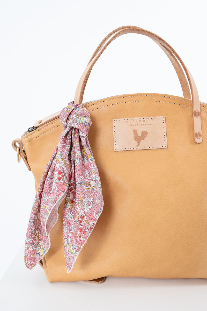 A tan leather handbag with a floral pink scarf tied to the handle, shown against a plain white background. The bag features a rectangular tan leather patch on the front with a rooster logo.  Handmade by Meanwhile Back on the Farm in Lynchburg,VA