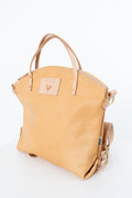 A light tan leather handbag with dual top handles, a zippered top, and a small rectangular logo patch on the front, photographed on a clean white background. Handmade by Meanwhile Back on the Farm in Lynchburg,VA