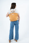 A woman viewed from behind wearing a tan leather backpack with tan straps, paired with a white ruffled top and wide-leg blue jeans, against a plain white studio background. Handmade by Meanwhile Back on the Farm in Lynchburg,VA