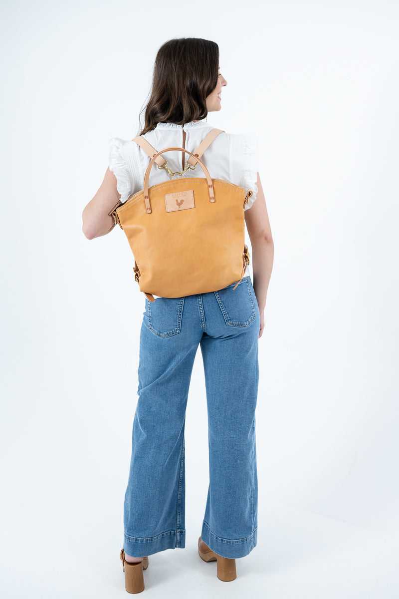 A woman viewed from behind wearing a tan leather backpack with tan straps, paired with a white ruffled top and wide-leg blue jeans, against a plain white studio background. Handmade by Meanwhile Back on the Farm in Lynchburg,VA