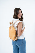 A woman with dark hair wearing a white ruffled top and blue jeans, shown from the back while wearing a light tan leather backpack against a plain white background. Handmade by Meanwhile Back on the Farm in Lynchburg,VA
