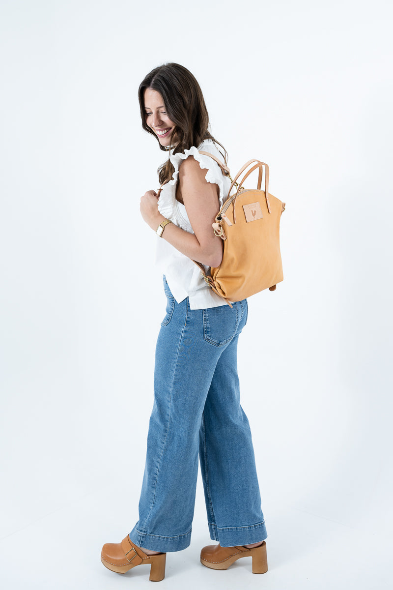 A woman wearing a white ruffled sleeveless top, wide-leg blue jeans, and brown platform heels, carrying a tan canvas backpack with gold-tone hardware against a plain white studio background. Handmade by Meanwhile Back on the Farm in Lynchburg,VA