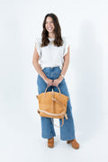 A woman stands against a white studio background wearing a white ruffled sleeveless top, blue wide-leg jeans, and brown boots, while holding a tan leather handbag with tan backpack straps. Handmade by Meanwhile Back on the Farm in Lynchburg,VA