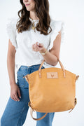 A woman wearing a white ruffled top and blue jeans holds a large mustard yellow leather tote bag with tan handles and a small rectangular logo patch on the front, photographed against a clean white background. Handmade by Meanwhile Back on the Farm in Lynchburg,VA