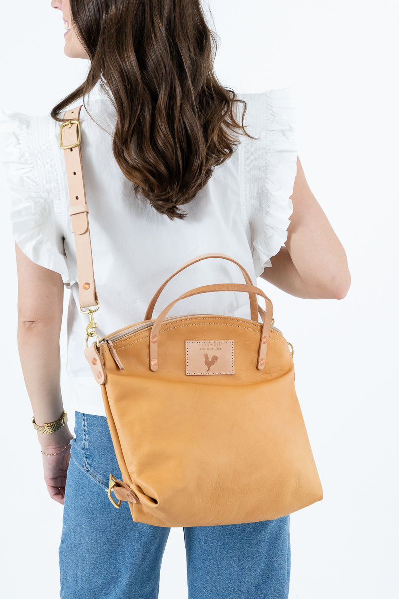 A woman wearing a white ruffled blouse and blue jeans is shown from the back, carrying a tan leather tote bag with a shoulder strap and top handles. The bag has a small rectangular logo patch on the front and gold-tone hardware. Handmade by Meanwhile Back on the Farm in Lynchburg,VA