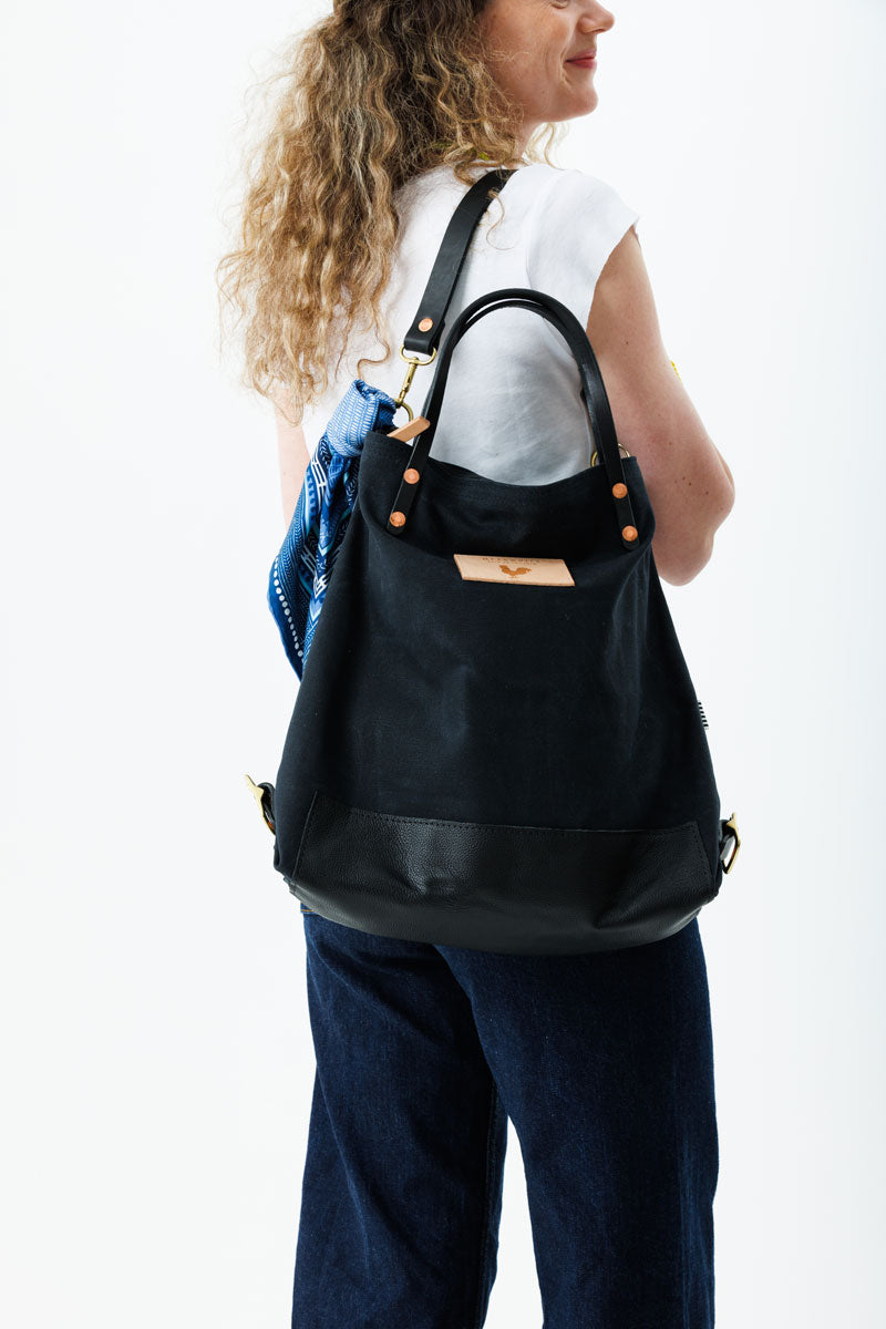 A woman wearing the black backpack with black straps and the meanwhile logo and a blue decorative scarf.