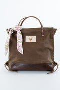 A brown canvas tote bag with dark brown leather trim, featuring a floral pink and white silk scarf tied to the side and a rectangular leather patch with a rooster logo on the front. Handmade by Meanwhile Back on the Farm in Lynchburg,VA
