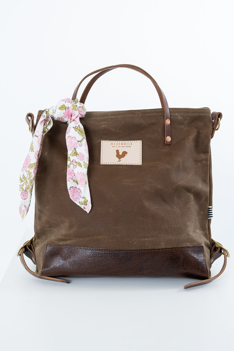 A brown canvas tote bag with dark brown leather trim, featuring a floral pink and white silk scarf tied to the side and a rectangular leather patch with a rooster logo on the front. Handmade by Meanwhile Back on the Farm in Lynchburg,VA