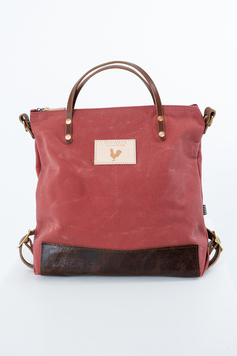 A red canvas tote bag with dark brown leather handles, a bottom panel, and hardware, featuring a small beige patch with a rooster logo on the front. Handmade by Meanwhile Back on the Farm in Lynchburg,VA