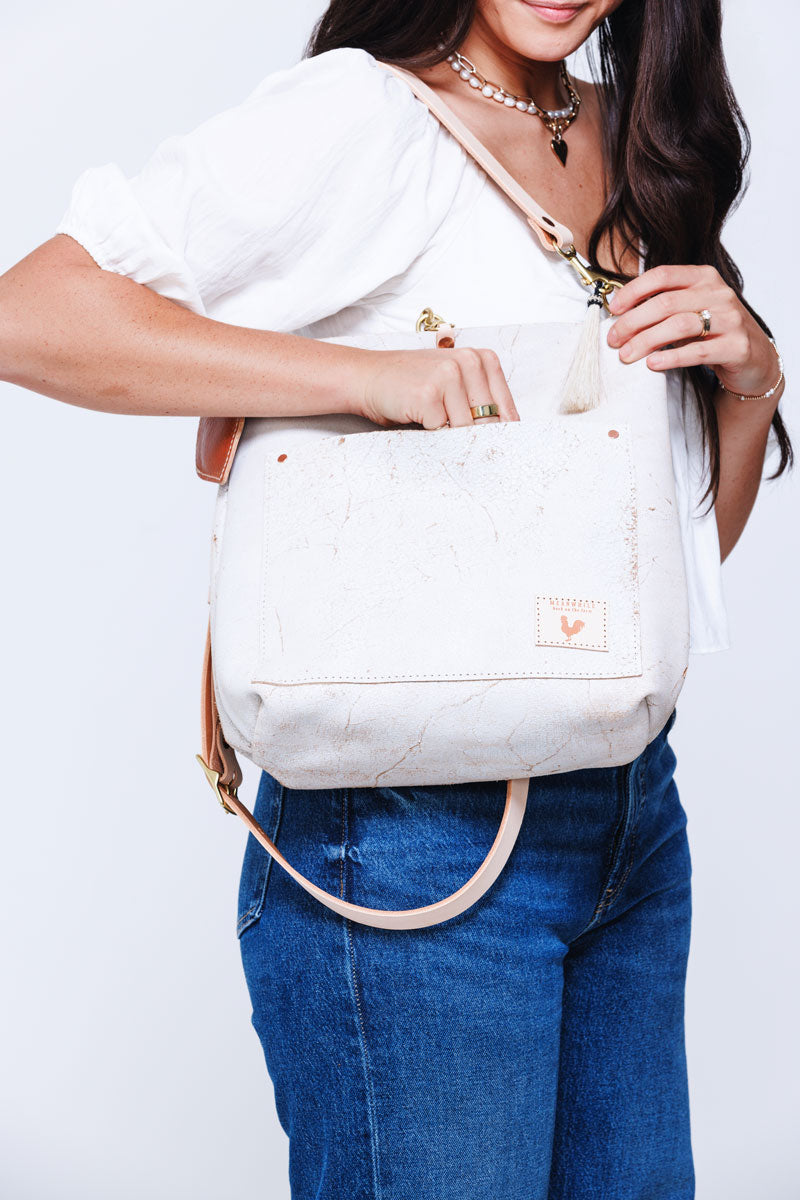 Picture of woman walking briskly in front of a wall carrying a large bundle of white hydrangeas that obscures her face.  She is wearing a large white handbag and cream colored handles and strap.