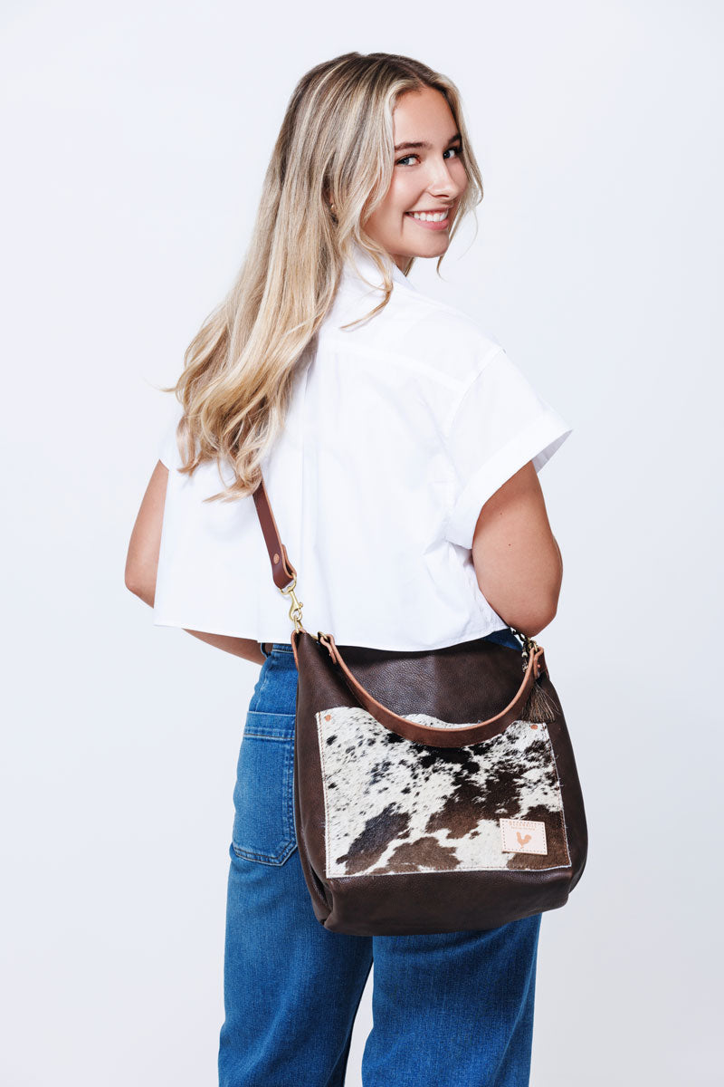 Picture of model holding a dark brown leather handbag in front of her. Bag has a front pocket made out of cowhide leather and dark brown handle strap.