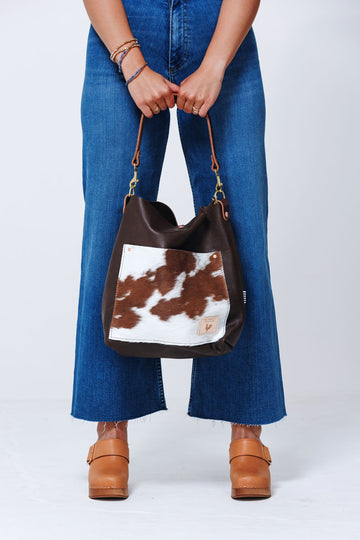 Picture of model holding a dark brown leather handbag in front of her. Bag has a front pocket made out of cowhide leather and dark brown handle strap.