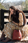 A woman sitting outdoors in front of a wooden fence, wearing a long-sleeve patterned dress, brown boots, and a dark brown fur vest. She is holding a reddish-brown leather bucket bag. Handcrafted by Meanwhile Back on the Farm in Lynchburg, Va.