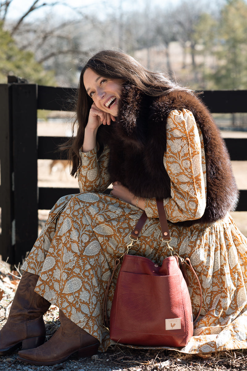 A woman sitting outdoors in front of a wooden fence, wearing a long-sleeve patterned dress, brown boots, and a dark brown fur vest. She is holding a reddish-brown leather bucket bag. Handcrafted by Meanwhile Back on the Farm in Lynchburg, Va.