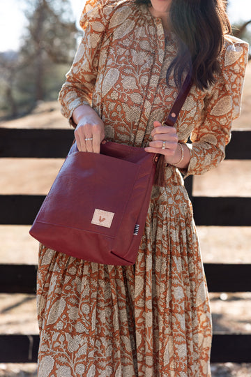 A woman wearing a rust-orange and beige paisley dress carries a burgundy leather shoulder bag with a small beige brand patch on the front, standing outdoors in front of a wooden fence. Handcrafted by Meanwhile Back on the Farm in Lynchburg, Va.