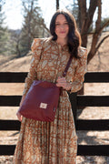 A woman wearing a long-sleeved beige and rust-orange patterned dress stands outdoors in front of a wooden fence, carrying a large burgundy leather shoulder bag. Handcrafted by Meanwhile Back on the Farm in Lynchburg, Va.