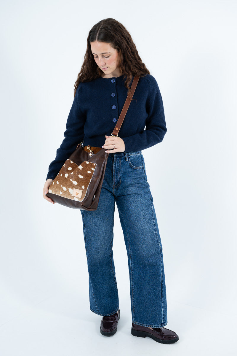A woman wearing a navy blue button-up sweater, wide-leg blue jeans, and dark brown leather shoes is shown from the waist down, holding a brown leather shoulder bag with a cowhide-patterned front panel, against a plain white studio background. Handcrafted in Lynchburg, VA.