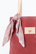 Close up of a red waxed canvas back with tan leather handles. There is a small flower scarf tied to the handles. Handmade by Meanwhile Back on the Farm in Lynchburg,VA