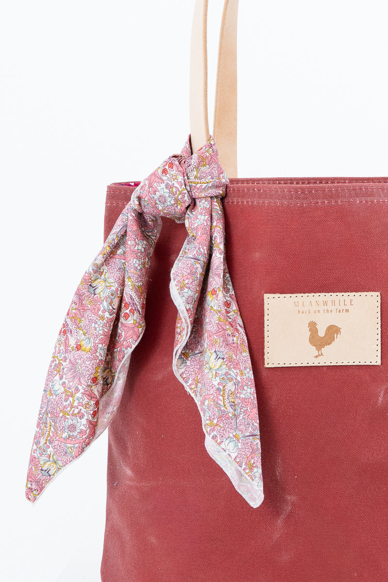 Close up of a red waxed canvas back with tan leather handles. There is a small flower scarf tied to the handles. Handmade by Meanwhile Back on the Farm in Lynchburg,VA