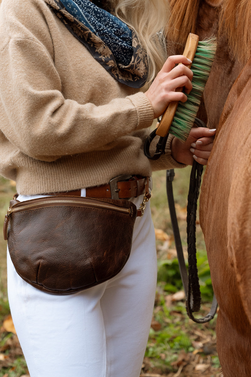 A woman wearing a white blouse and blue jeans models a brown leather crescent-shaped belt bag (fanny pack) with a gold zipper, set against a plain white studio background. Handmade by Meanwhile Back on the Farm in Lynchburg,VA