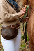 A person in a beige sweater and white pants holds a brown leather fanny pack and a horse brush next to a brown horse outdoors. Handcrafted by Meanwhile Back on the Farm in Lynchburg, Va.