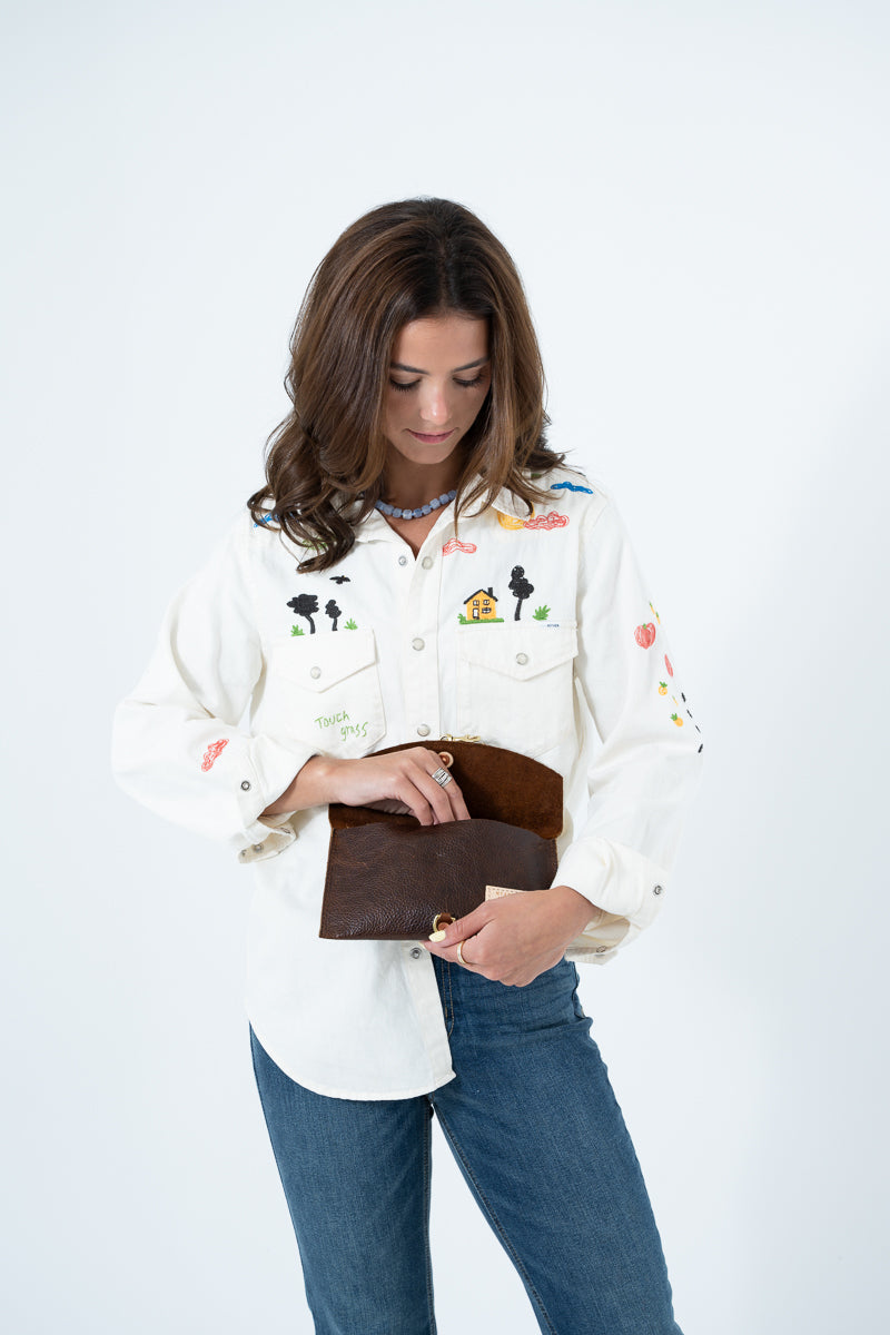 A woman wearing a cream-colored embroidered button-up shirt and blue jeans, holding a brown leather clutch purse against a plain white studio background. Handmade by Meanwhile Back on the Farm in Lynchburg,VA