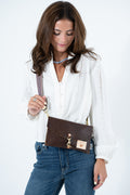 A woman wearing a white eyelet blouse and blue jeans carries a brown leather crossbody bag with gold hardware and a small tan logo patch, photographed against a clean white studio background. Handmade by Meanwhile Back on the Farm in Lynchburg,VA