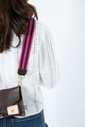 A close-up lifestyle shot of a model wearing a white eyelet blouse and blue jeans, carrying a dark brown textured leather crossbody bag with a vibrant pink and purple striped woven strap. Handmade by Meanwhile Back on the Farm in Lynchburg,VA