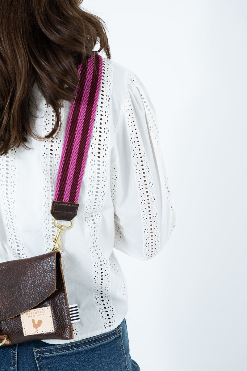 A close-up lifestyle shot of a model wearing a white eyelet blouse and blue jeans, carrying a dark brown textured leather crossbody bag with a vibrant pink and purple striped woven strap. Handmade by Meanwhile Back on the Farm in Lynchburg,VA