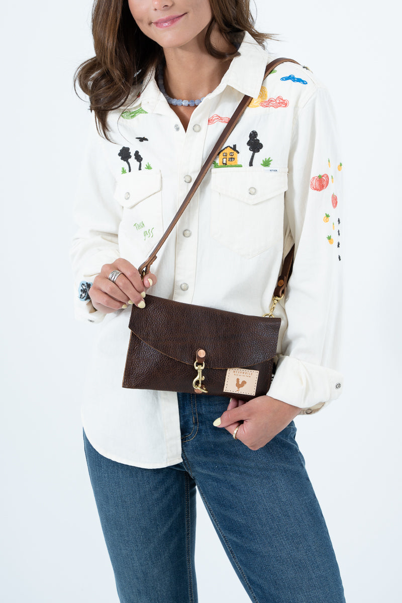 A woman wearing a white embroidered button-up shirt and blue jeans carries a brown leather shoulder bag with a brown strap and gold-tone hardware against a plain white background. Handmade by Meanwhile Back on the Farm in Lynchburg,VA