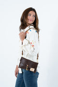A woman wearing a white embroidered shirt and blue jeans models a brown leather crossbody bag with gold hardware against a plain white background. Handmade by Meanwhile Back on the Farm in Lynchburg,VA