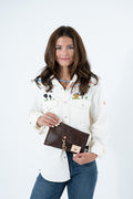A woman modeling a white embroidered button-up shirt and blue jeans, holding a brown leather clutch bag with a gold clasp. Handmade by Meanwhile Back on the Farm in Lynchburg,VA