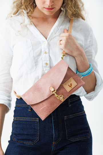 Woman wearing the rose pink colored bag with a hook that closes the bag, meanwhile logo, and a light brown cross body strap.