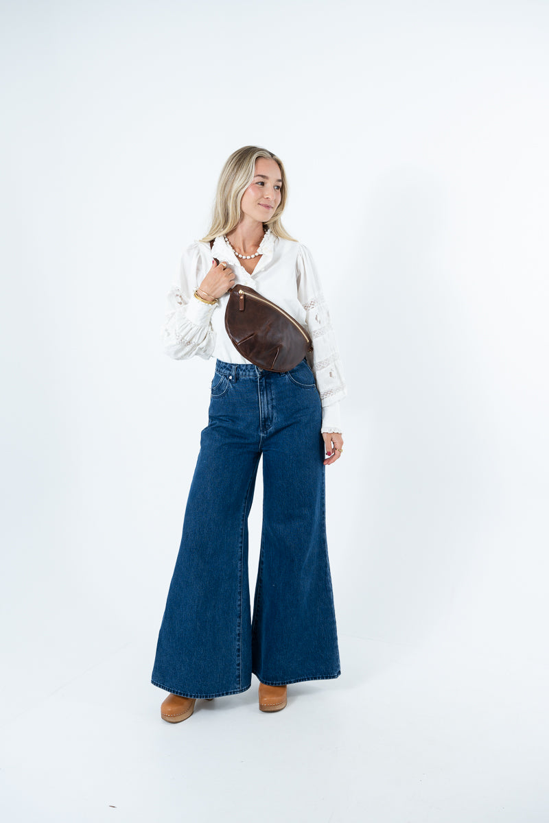 A woman modeling a white long-sleeve button-up shirt, dark blue wide-leg denim jeans, and brown leather shoes, carrying a brown leather crossbody bag against a plain white studio background. Handmade by Meanwhile Back on the Farm in Lynchburg,VA
