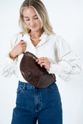 A woman wearing a white blouse and blue jeans models a dark brown leather crescent-shaped shoulder bag with a gold-tone zipper. Handmade by Meanwhile Back on the Farm in Lynchburg,VA