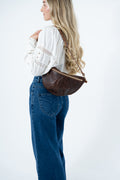 A woman wearing a white blouse and blue jeans is shown from the back and side, carrying a dark brown crescent-shaped leather shoulder bag against a plain white studio background. Handmade by Meanwhile Back on the Farm in Lynchburg,VA