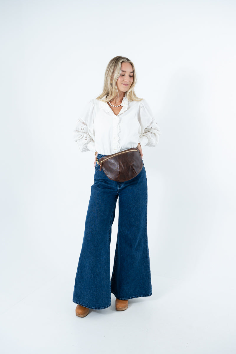 A woman modeling a white long-sleeve blouse, dark blue wide-leg denim jeans, and a brown leather fanny pack against a plain white studio background. Handmade by Meanwhile Back on the Farm in Lynchburg,VA