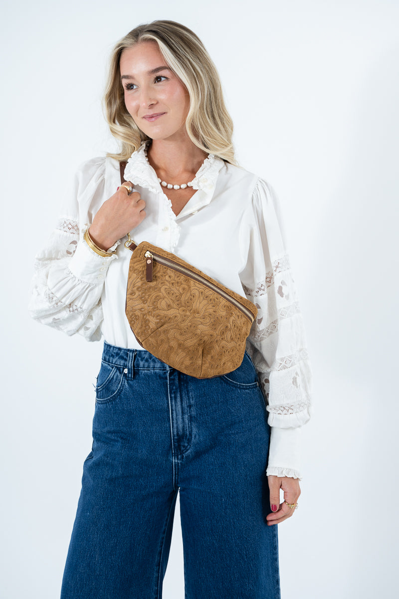 A woman wearing a white lace-sleeve blouse and blue jeans models a tan brown textured leather fanny pack with a gold zipper, set against a plain white studio background. Handmade by Meanwhile Back on the Farm in Lynchburg,VA