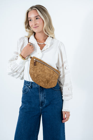 A woman wearing a white lace-sleeve blouse and blue jeans models a tan brown textured leather fanny pack with a gold zipper, set against a plain white studio background. Handmade by Meanwhile Back on the Farm in Lynchburg,VA