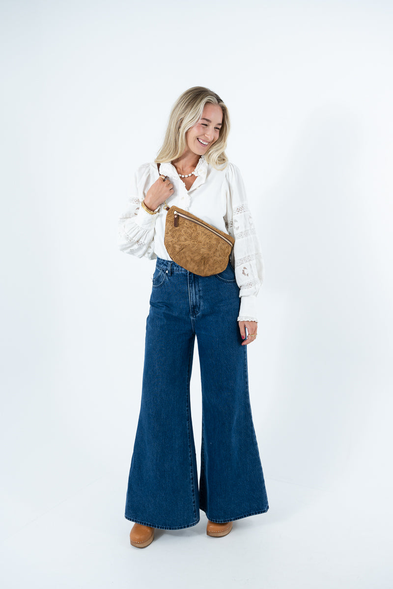 A woman modeling a white long-sleeve blouse, dark blue wide-leg denim pants, and a tan brown suede crossbody bag against a plain white studio background. Handmade by Meanwhile Back on the Farm in Lynchburg,VA