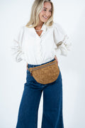 A woman wearing a white blouse with lace sleeves, blue wide-leg jeans, and a brown tan leather fanny pack, standing against a plain white background. Handmade by Meanwhile Back on the Farm in Lynchburg,VA