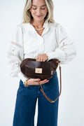A woman wearing a white lace-sleeve blouse and blue jeans holds a brown leather crossbody bag with a small rectangular logo patch. The photo is taken against a clean white studio background. Handmade by Meanwhile Back on the Farm in Lynchburg,VA