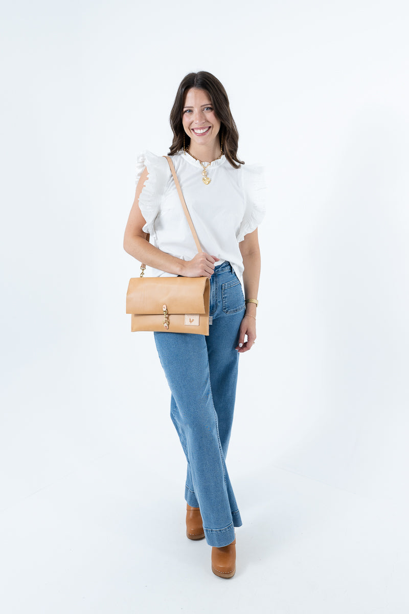 A woman modeling a light tan leather fold over clutch bag with gold hardware, styled with a white ruffled top, blue jeans, and brown boots against a white studio background. Handmade by Meanwhile Back on the Farm in Lynchburg,VA