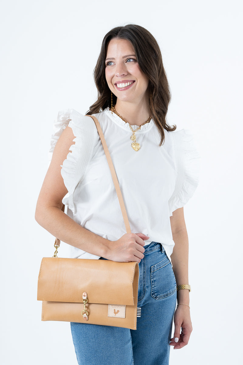 A woman wearing a white ruffled-sleeve blouse and blue jeans carries a tan leather crossbody bag with gold hardware against a plain white studio background. Handmade by Meanwhile Back on the Farm in Lynchburg,VA