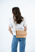 A woman shown from behind wearing a white ruffled-sleeve top and blue jeans, carrying a tan leather crossbody bag with a gold-tone clasp and strap against a plain white background. Handmade by Meanwhile Back on the Farm in Lynchburg,VA