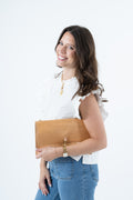 A woman with long brown hair smiling and looking over her shoulder, wearing a white ruffled top and blue jeans while holding a tan leather clutch bag against a plain white background.Handmade by Meanwhile Back on the Farm in Lynchburg,VA